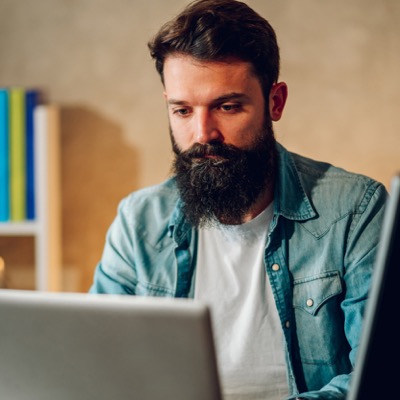 Man with a beard focused on working at a laptop in a home or office setting.