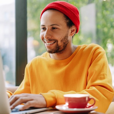 Young man wearing a red beanie and orange sweater sitting at a table with a laptop and a cup of coffee.